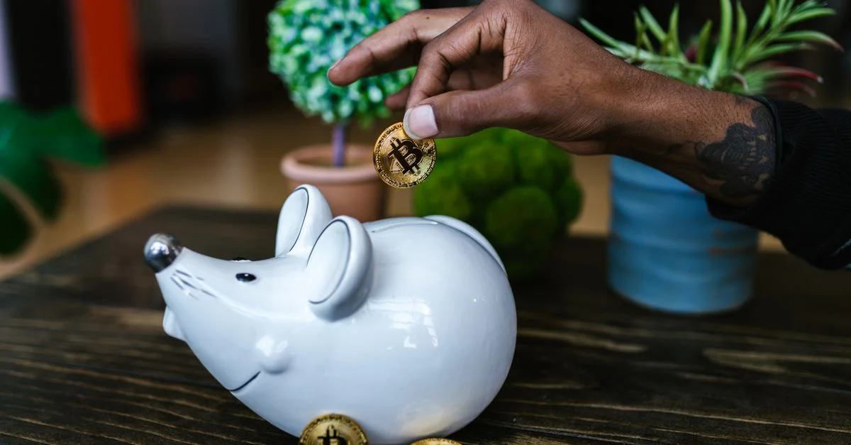 Gros plan sur une main plaçant un bitcoin dans une tirelire en forme de souris sur une table.