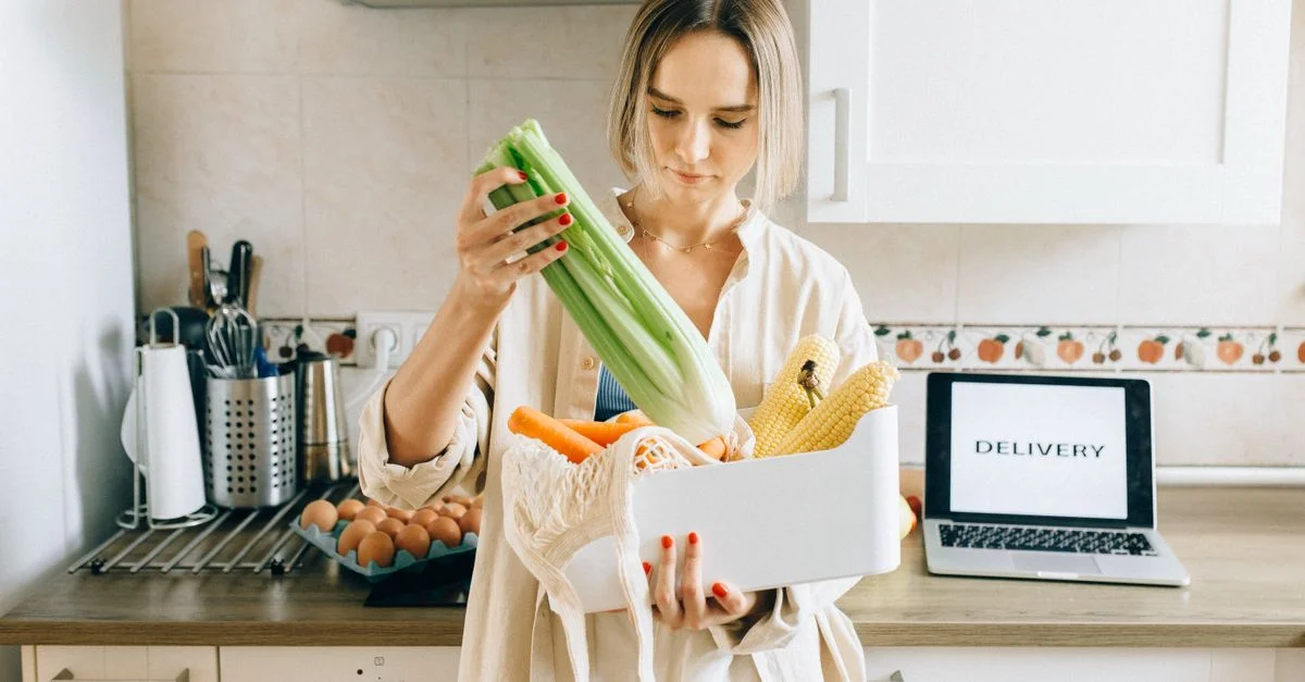 Femme déballant des légumes frais d'une boîte de livraison dans une cuisine moderne avec un ordinateur portable à proximité.